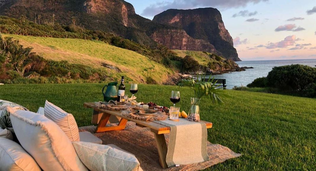 A romantic picnic setup on a green lawn with snacks and wine, overlooking the ocean and cliffs at sunset.