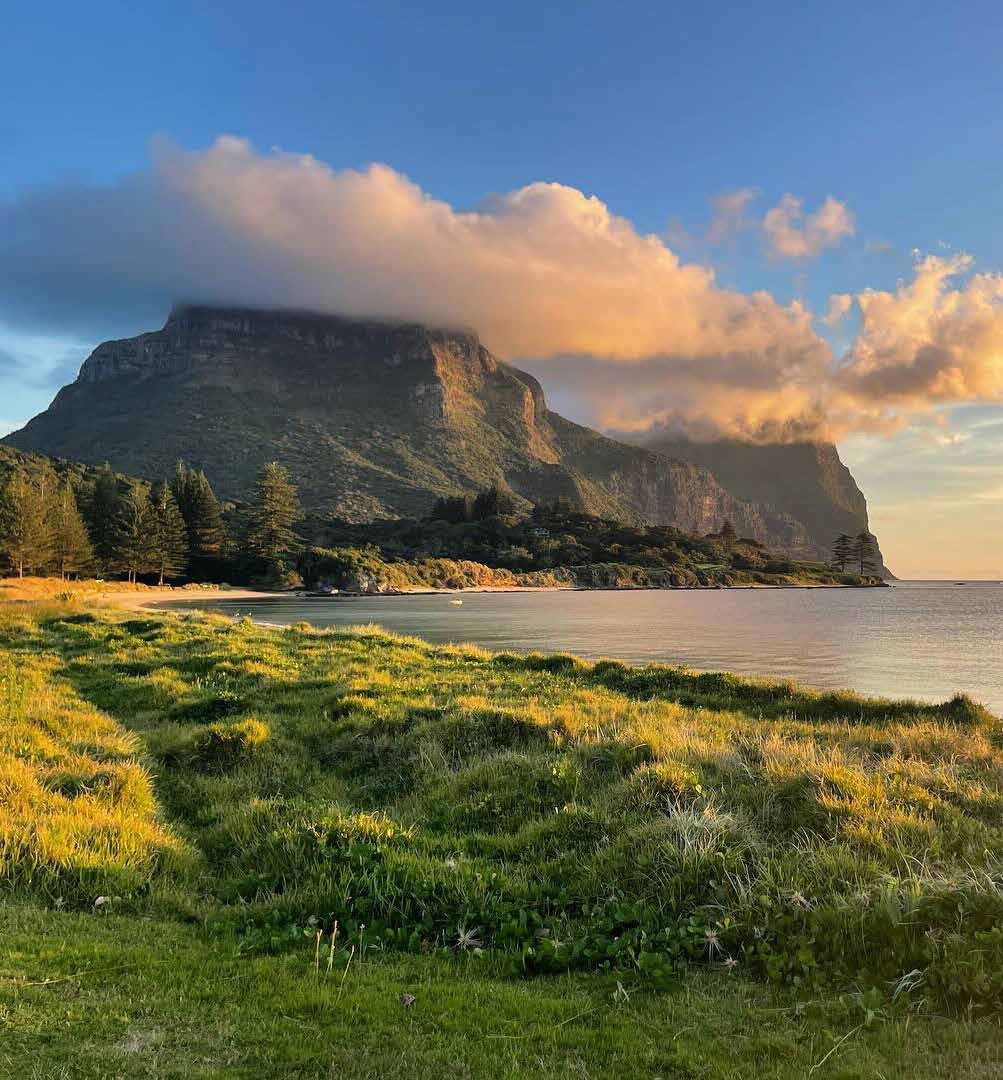 A small mountain with clouds covering the top, green trees a beach, grassed land and the ocean in the foreground.