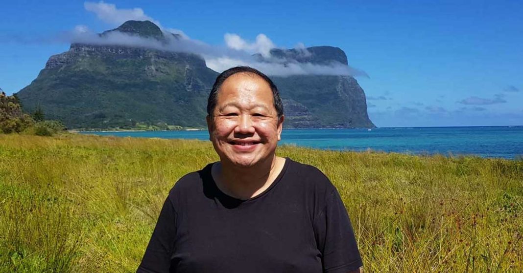 Stephen Sia, owner of the Lord Howe Island Signal, standing in tall grasses, smiling into the camera with a mountain and lagoon behind him.