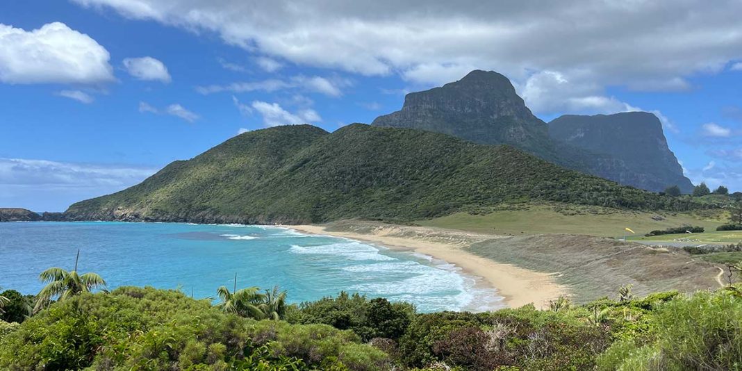 An idyllic coastal scene showcasing a broad view of a blue lagoon with coral patches, a sandy shore, lush green hills, and two towering, rugged mountain peaks under a cloudy blue sky.