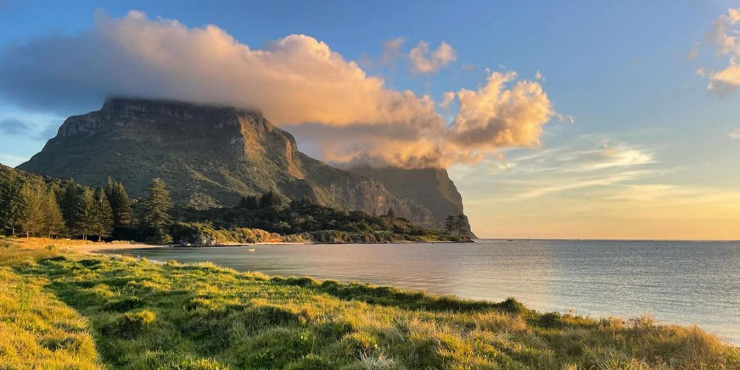 This image is a gorgeous beach scene. In the foreground, there is a lush green shoreline. In the background, a tall mountain sits under soft clouds.