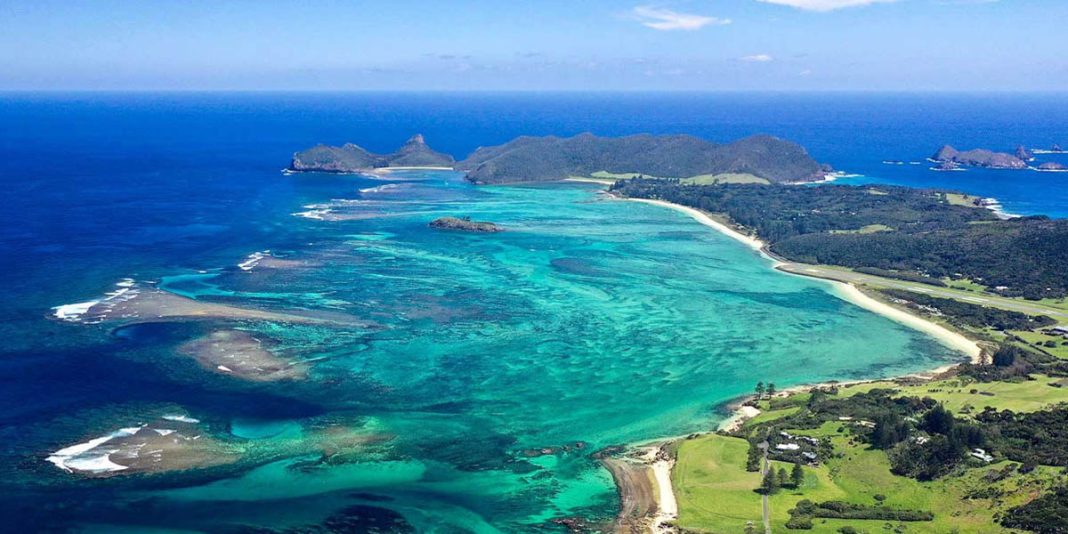 An aerial view of a lush coastal landscape with vibrant turquoise waters, reef formations, sandy beaches, and a series of green, hilly islets stretching into the distance under a clear blue sky.