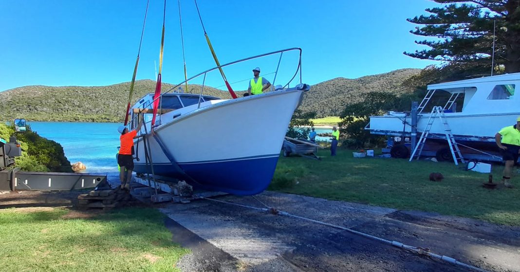 A boat being hoisted by a crane on a bright day, with workers assisting in the operation near a calm bay and green hills in the background.