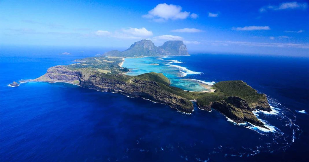 An aerial view of Lord Howe Island, featuring its distinctive twin peaks, lush green terrain, surrounding coral reef lagoons, and the vast blue ocean.