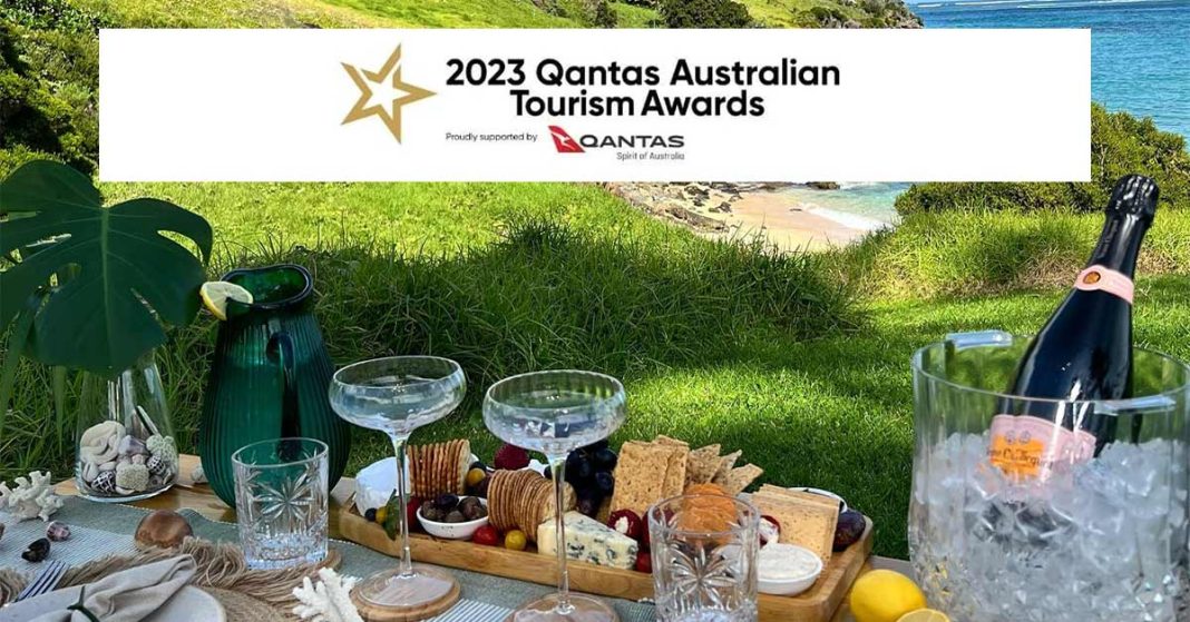 A banner image for the 2023 Qantas Australian Tourism Awards, featuring a luxurious picnic setup on a grassy knoll overlooking a secluded beach. The picnic includes a variety of gourmet snacks, a bottle of champagne on ice, and elegant glassware, set against a backdrop of lush coastal vegetation.