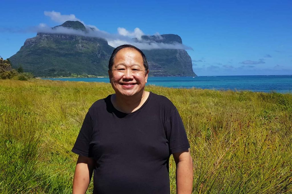 Stephen Sia, owner of the Lord Howe Island Signal, standing in tall grasses, smiling into the camera with a mountain and lagoon behind him.