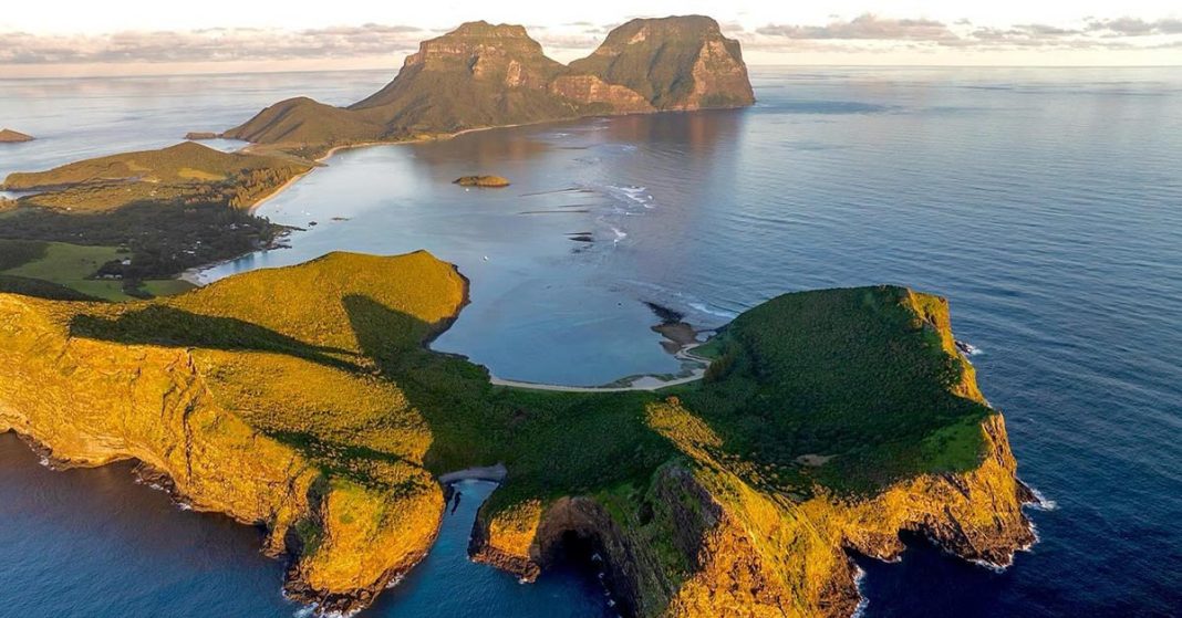 This aerial view image captures the stunning landscape of Lord Howe Island, featuring the prominent Mount Lidgbird and Mount Gower in the background. The Island's lush greenery, pristine beaches, and crystal-clear waters create a picturesque and serene environment.