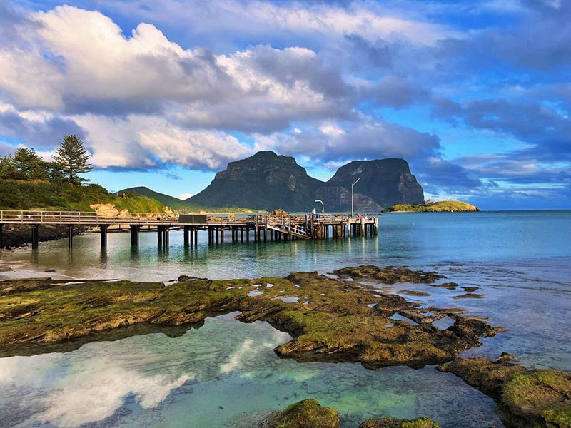 This image captures the stunning landscape of Lord Howe Island, featuring the prominent Mount Lidgbird and Mount Gower in the background and the jetty in the foreground. The Island's lush greenery, pristine beaches, and crystal-clear waters create a picturesque and serene environment.