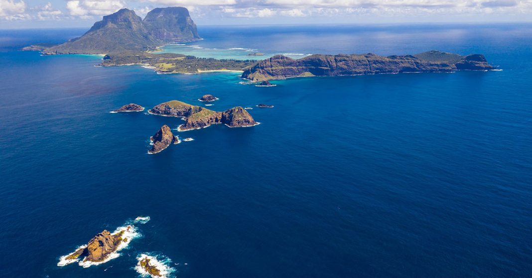 This aerial image captures the stunning Lord Howe Island, featuring the prominent Mount Lidgbird and Mount Gower in the background. The Island's lush greenery, pristine beaches, and crystal-clear waters create a picturesque and serene environment.